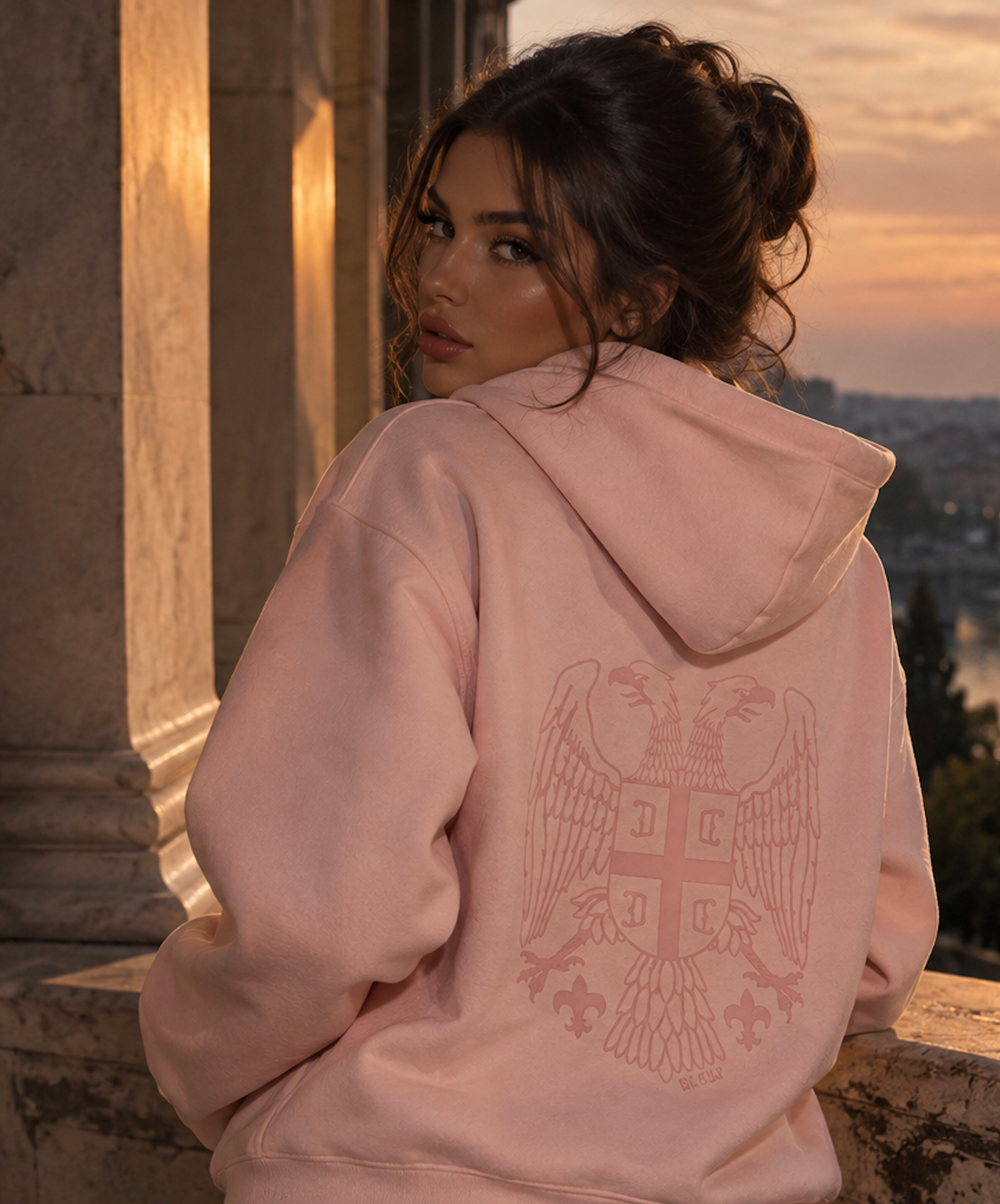 A woman wearing a light pink Serbian hoodie with a large tonal Srpski crest graphic on the back, photographed outdoors against a warm sunset and stone columns.
